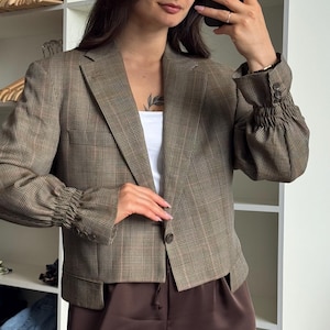 Könnte beinhalten: Ein brauner Blazer mit Karomuster, fallendem Revers, einem Knopfverschluss vorne und gerafften, elastischen Ärmelbündchen mit jeweils drei Zierknöpfen. Die kurz geschnittene Jacke wird über einem weißen Oberteil und einer dunkelbraunen Hose getragen.