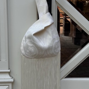 Può includere: Una borsa da polso bianca caratterizzata da un sottile motivo floreale damascato tono su tono e da una lunga frangia dritta lungo il bordo inferiore. La borsa presenta un unico manico integrato annodato in alto, creando una silhouette morbida.