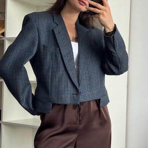 Pode incluir: Um blazer curto com padrão pied-de-poule azul-escuro e preto, mangas compridas, lapela entalhada e fecho frontal com um botão. O blazer é combinado com calças de cetim castanho-escuro com cintura de cordão e corte descontraído.