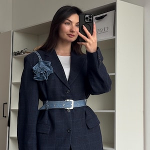 Pode incluir: Um blazer azul escuro com cinto de ganga e uma flor decorativa de ganga no ombro. O blazer tem um padrão xadrez e é combinado com calças pretas. O cinto tem uma fivela prateada.