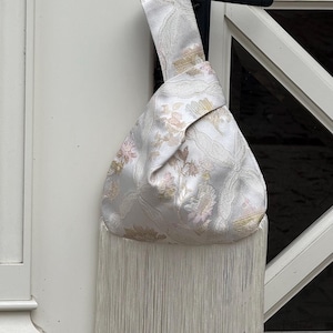 May include: A white wristlet bag featuring a subtle floral jacquard pattern in soft pink and gold tones. The bag has a knotted handle design and is finished with a long, straight white fringe trim that hangs from the bottom edge of the pouch.