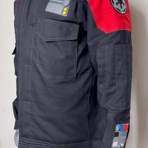 Pode incluir: Casaco preto com detalhes vermelhos nos ombros e design acolchoado. Possui v&aacute;rios bolsos, patches e um emblema circular no ombro. O casaco tem gola alta e &eacute; exibido em um manequim.