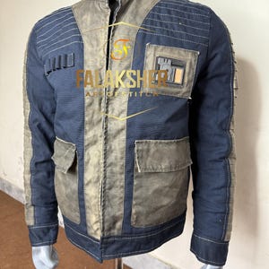 Puede incluir: Chaqueta azul marino y gris con cuello alto y múltiples bolsillos. La chaqueta presenta un parche en el pecho y costuras decorativas. El texto "FalakSher Art of Stitch" es visible en la chaqueta.