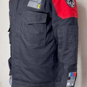 Pode incluir: Casaco preto com detalhes vermelhos nos ombros, com v&aacute;rios bolsos e patches. O casaco tem um design de ombro vermelho acolchoado e um patch com um emblema circular. O casaco &eacute; exibido em um manequim.