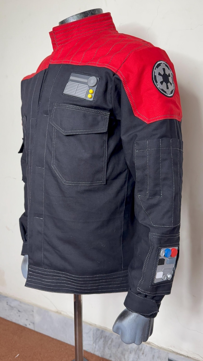 Pode incluir: Casaco preto com detalhes vermelhos nos ombros, com v&aacute;rios bolsos e patches. O casaco tem um design de ombro vermelho acolchoado e um patch com um emblema circular. O casaco &eacute; exibido em um manequim.