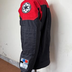 Pode incluir: Casaco preto e vermelho com um emblema circular branco e preto no ombro. O casaco tem uma gola alta e v&aacute;rios bolsos. A manga apresenta um design de painel de controle com detalhes azuis, vermelhos e prateados.