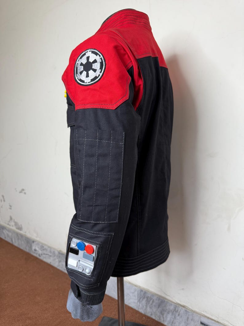 Pode incluir: Casaco preto e vermelho com um emblema circular branco e preto no ombro. O casaco tem uma gola alta e v&aacute;rios bolsos. A manga apresenta um design de painel de controle com detalhes azuis, vermelhos e prateados.