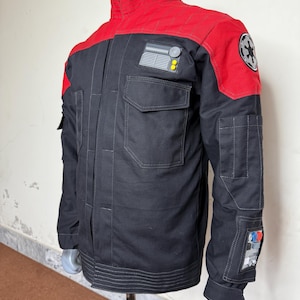 以下が含まれることがあります： ハイカラーと複数のポケットが付いた黒と赤のジャケット。ジャケットは赤い肩ヨークと袖にパッチが付いています。円形のエンブレムが右肩にあります。ジャケットはマネキンに表示されています。