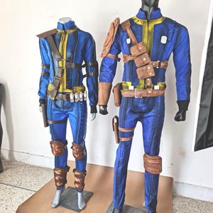 May include: Two mannequins wearing blue jumpsuits with brown leather accents and straps. The jumpsuits have yellow accents and are styled with knee pads and holsters.