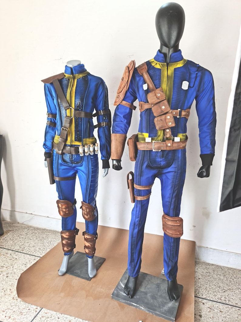 May include: Two mannequins wearing blue jumpsuits with brown leather accents and straps. The jumpsuits have yellow accents and are styled with knee pads and holsters.