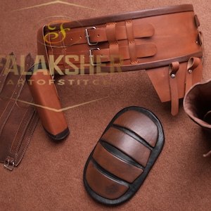 May include: A collection of brown leather armor pieces, including a wide belt with buckles, a leg guard, a knee pad, and a quiver. The pieces are arranged on a brown surface. The text "FALAKSHER ARTOFSTITCH" is visible.