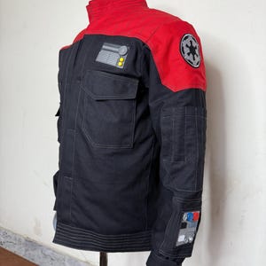 Pode incluir: Casaco preto e vermelho com v&aacute;rios bolsos e patches. O casaco apresenta uma gola alta e um patch no ombro com um emblema circular. Patches adicionais com designs gr&aacute;ficos est&atilde;o nas mangas. O casaco &eacute; exibido em um manequim.