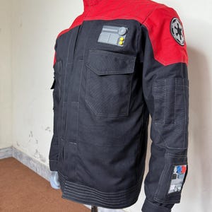 Pode incluir: Casaco preto e vermelho com gola alta e v&aacute;rios bolsos. O casaco apresenta um patch no ombro com um emblema circular e outro patch no peito com um design retangular. As mangas t&ecirc;m detalhes adicionais.