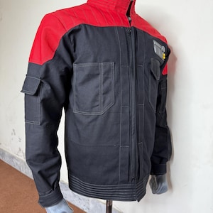 Pode incluir: Casaco preto e vermelho com gola alta e fecho de correr. O casaco apresenta dois bolsos frontais, dois bolsos no peito e um pequeno remendo na parte superior direita do peito. Ombros e parte superior das costas acolchoados em vermelho.
