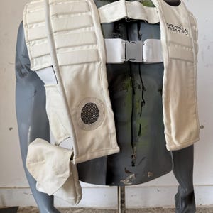 May include: Cream-colored tactical vest with multiple padded sections and a mesh vent. The vest features adjustable white straps and a silver buckle closure. A small pouch is attached to the side. The vest is displayed on a mannequin.