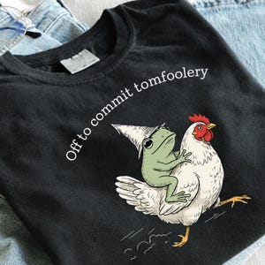 May include: A black t-shirt featuring a graphic of a light green frog wearing a pointed hat while riding on the back of a white chicken. The text "Off to commit tomfoolery" is printed in white above the illustration in a simple sans-serif font.