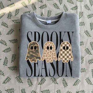 May include: A heather gray crewneck sweatshirt featuring the black text SPOOKY SEASON. Three ghost-shaped appliques are centered on the front, each with a different pattern: one in green camouflage, one in tan leopard print, and one in a brown checkerboard.