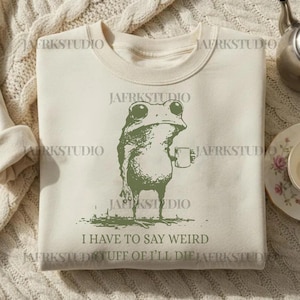 Op de afbeelding: Crèmekleurige sweatshirt met een groene kikker opdruk die een mok vasthoudt. De tekst "I HAVE TO SAY WEIRD STUFF OF I'LL DIE" staat eronder. De sweatshirt heeft een ronde hals en lange mouwen.