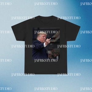 May include: Black t-shirt featuring a photographic print of a man in a suit, raising his middle finger. The image is centered on the shirt. The text "JAFRKSTUDIO" is printed multiple times around the shirt.