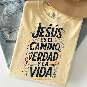 May include: A light yellow t-shirt with the text "Jesús es el camino, la verdad y la vida" in navy blue, with colorful accents. The shirt is folded on top of blue jeans and a beige hat.