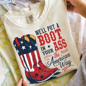 May include: A cream-colored t-shirt featuring a graphic of two cowboy boots with a red, white, and blue American flag pattern. The text reads: WE'LL PUT A BOOT IN YOUR ASS IT'S THE AMERICAN WAY, printed in bold red and dark blue lettering.