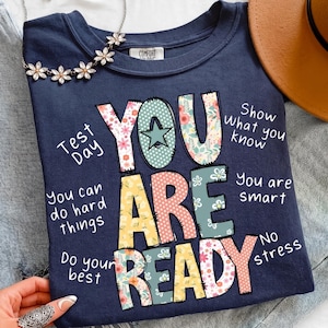 Op de afbeelding: Marineblauw T-shirt met de woorden "YOU ARE READY" in kleurrijke, bloemmotief letters. Extra tekst bevat "Test Day", "You can do hard things" en "Show what you know". Een bloemenketting en een bruine hoed zijn ook zichtbaar.
