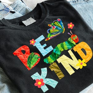 May include: A black t-shirt featuring the text BE KIND in colourful, textured letters. The design includes a green caterpillar, a multi-coloured butterfly, and small red and yellow flowers. The shirt is layered over light blue denim fabric.
