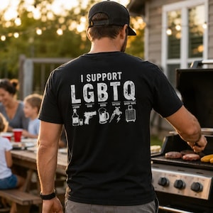 May include: A black t-shirt featuring the white text I SUPPORT LGBTQ with icons representing liquor, guns, beers, tattoos, and quick trips. The design is printed on the back of the shirt, which is worn by an individual standing near a grill.