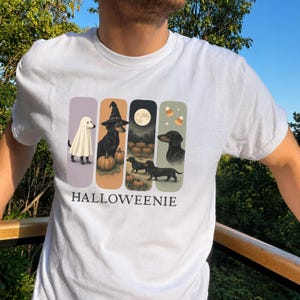 Puede incluir: Una camiseta blanca con un gráfico de cuatro paneles verticales que muestran perros salchicha con disfraces de Halloween, calabazas, una luna llena y dulces. El texto HALLOWEENIE está impreso en letras mayúsculas negras bajo el diseño.