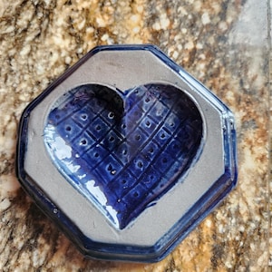 May include: A handmade ceramic dish with a heart-shaped indentation. The dish is a deep blue color with a textured pattern inside the heart and a gray border. The dish has an octagonal shape.
