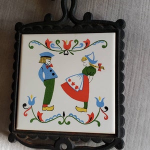 May include: A vintage trivet with a black wrought iron frame. The white ceramic tile insert features a colorful illustration of two Dutch children in traditional clothing. The design includes red, blue, green, and yellow accents, with floral motifs.