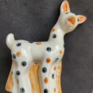 May include: A ceramic figurine of a deer with a white body, orange and black spots, and orange accents on the ears and legs. The deer has a slightly open mouth and is standing upright.