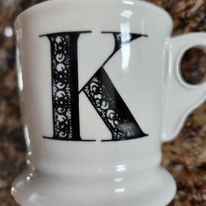 May include: White ceramic mug with a black letter "K" on the front. The letter is decorated with a detailed, ornate pattern. The mug has a rounded base and a handle on the side. A simple and elegant design for a coffee or tea cup.