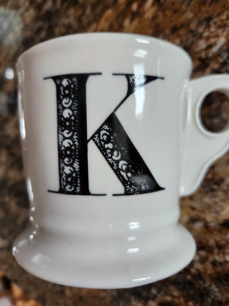 May include: White ceramic mug with a black letter "K" on the front. The letter is decorated with a detailed, ornate pattern. The mug has a rounded base and a handle on the side. A simple and elegant design for a coffee or tea cup.