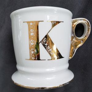May include: White ceramic mug with a gold-colored handle and the letter "K" in gold. The letter has an ornate design. The rim and base of the mug are also trimmed in gold.
