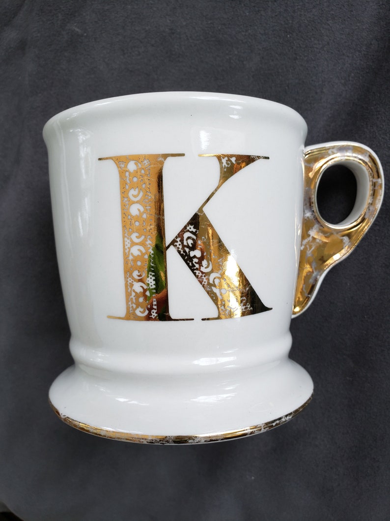 May include: White ceramic mug with a gold-colored handle and the letter "K" in gold. The letter has an ornate design. The rim and base of the mug are also trimmed in gold.