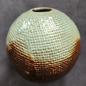 May include: A spherical ceramic vase with a textured woven pattern. The top half is a light teal color, transitioning to a rich brown at the bottom. The vase has a small opening at the top.