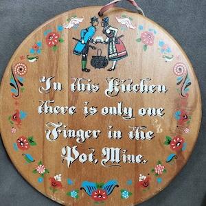 May include: Round wooden kitchen sign with a decorative border of red, blue, and white floral designs. The sign features a couple in traditional attire and the text "In this Kitchen there is only one Finger in the Pot, Mine."