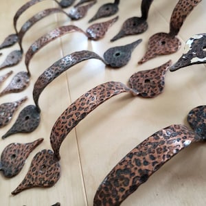 May include: Close-up of several hammered copper drawer pulls and matching backplates. The pulls have a textured, speckled surface and a curved handle design. The backplates are teardrop-shaped with a single hole.