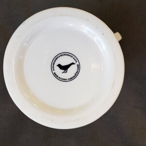 May include: A white ceramic mug base with a small handle. The bottom of the mug features a black bird silhouette inside a circular design with the text "Dishwasher & Microwave Safe" and "Made in China".