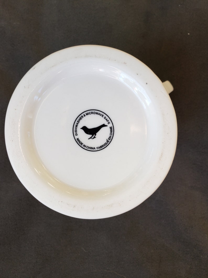 May include: A white ceramic mug base with a small handle. The bottom of the mug features a black bird silhouette inside a circular design with the text "Dishwasher & Microwave Safe" and "Made in China".