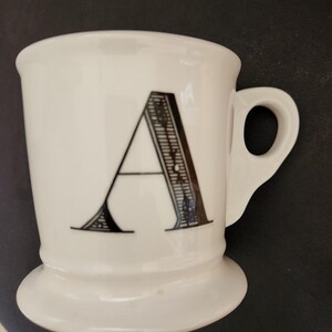 May include: White ceramic mug with a black letter "A" in a decorative font. The mug has a rounded base and a handle. The mug is likely used for drinking coffee, tea, or other beverages.