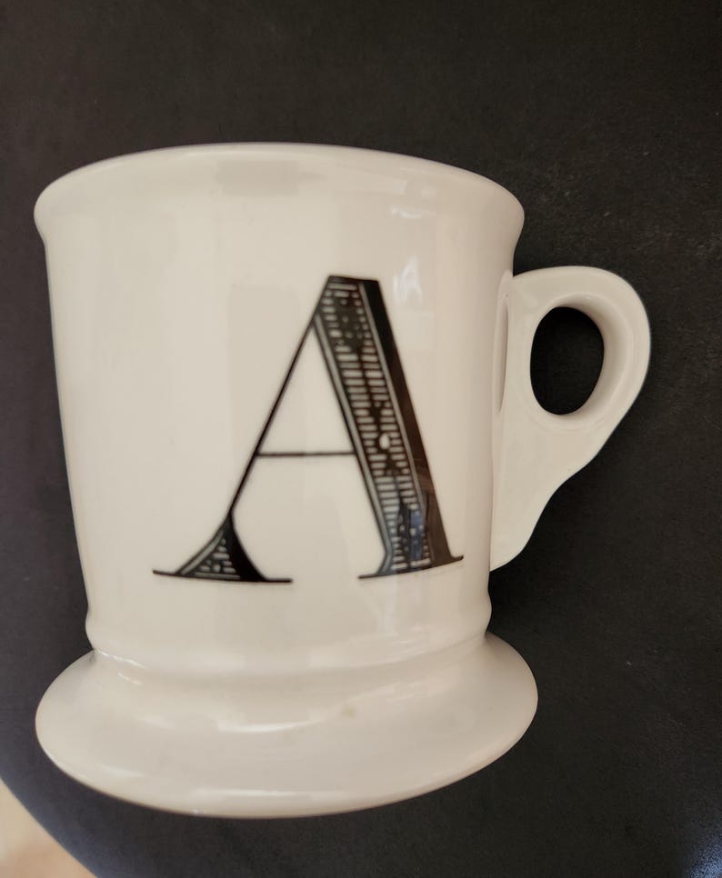 May include: White ceramic mug with a black letter "A" in a decorative font. The mug has a rounded base and a handle. The mug is likely used for drinking coffee, tea, or other beverages.