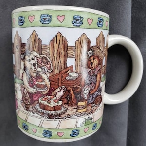 May include: White ceramic mug with a whimsical illustration of bears and a rabbit having a picnic. The design includes a wooden fence, cakes, and a honey jar. The rim features a green border with hearts and teacups.