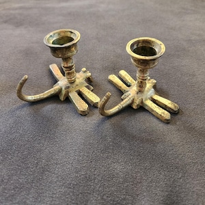 May include: Two antique brass candle holders. Each holder has a circular cup for the candle and a dragonfly-shaped base with curved hooks. The brass has a tarnished, aged appearance, suggesting they are vintage or antique.