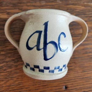 May include: A white ceramic pitcher with two handles and a blue and white checkered pattern around the bottom. The pitcher has the letters "abc" written in blue on the front.