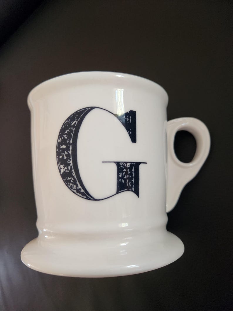 May include: White ceramic mug with a large, ornate black letter "G" on the front. The mug has a handle and a wide, stable base. The design is simple yet elegant, suitable for coffee or tea.