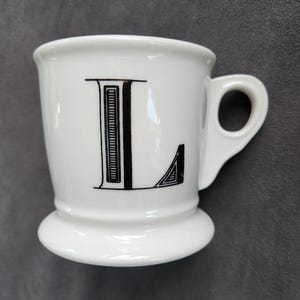 May include: White ceramic mug with a black letter "L" on the front. The mug has a rounded base and a small handle. The letter "L" is in a bold, decorative font.