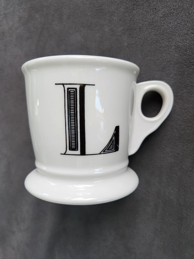 May include: White ceramic mug with a black letter "L" on the front. The mug has a rounded base and a small handle. The letter "L" is in a bold, decorative font.