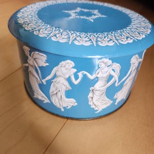 May include: A round, light blue decorative tin with a white relief design. The lid features a white star and floral border. The sides depict white classical figures in a dancing pose. A vintage-style storage container.